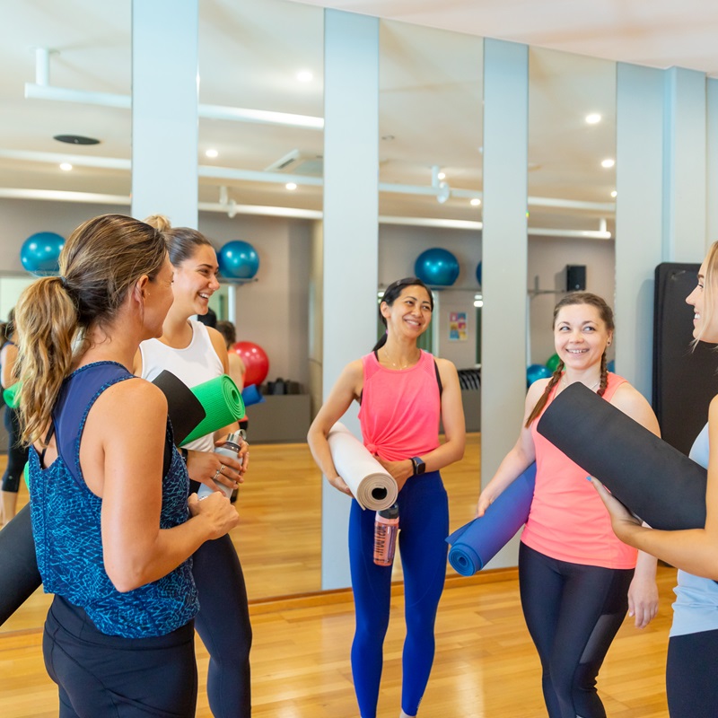 A photo of five women in active wear holding yoga mats talking amongst themselves