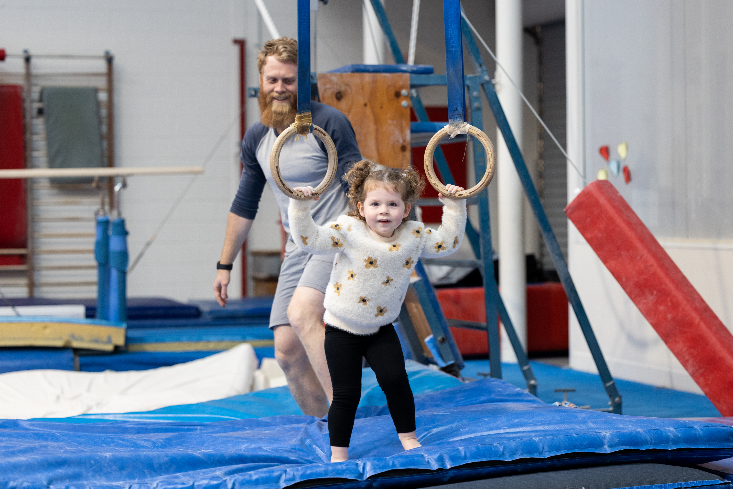 A child holding onto a pair of gymnastics stationary rings as her dad looks on at the Perth HPC Kids Gymnastics in Mount Claremont, Perth
