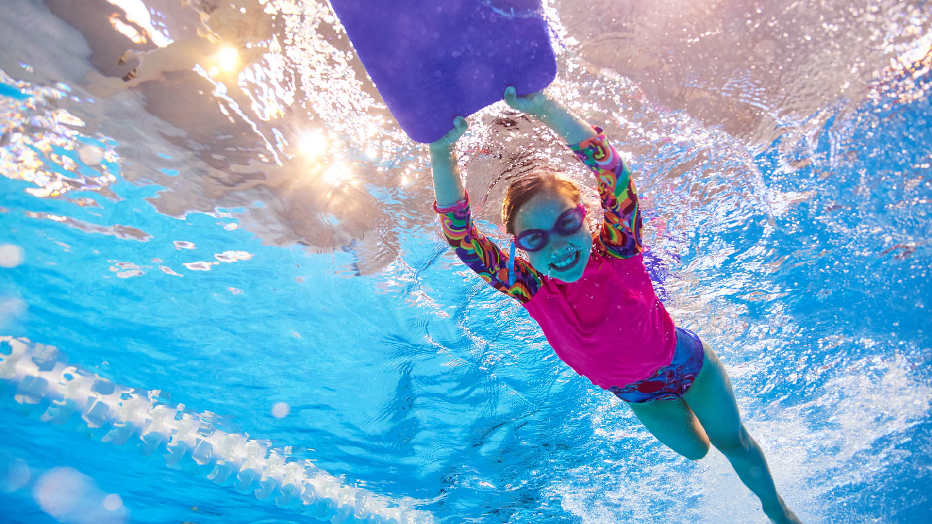 An underwater photo of a girl smiling as she swims along whilst holding at kick board at Perth HPC in Mount Claremont